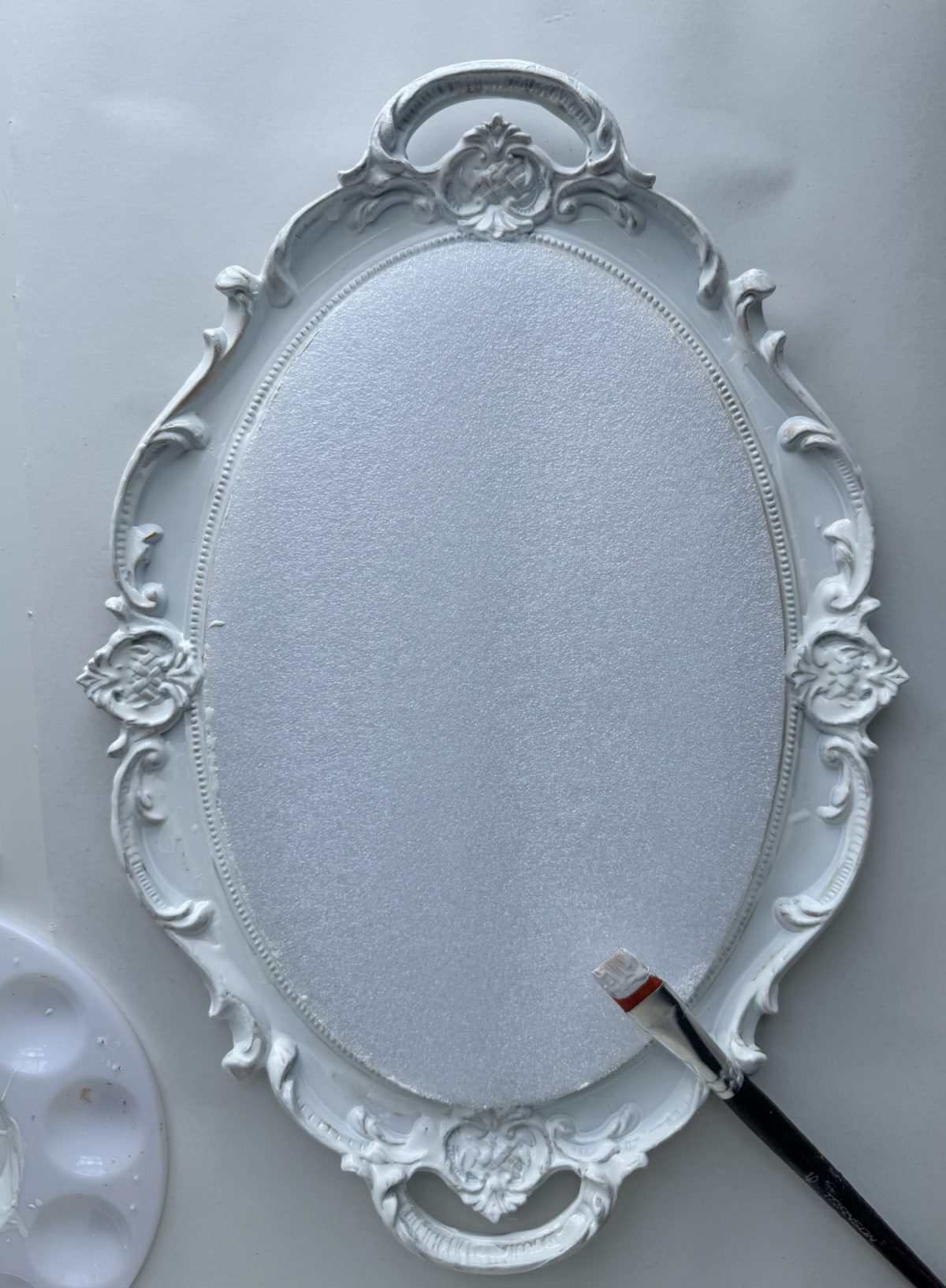 An ornate, empty oval frame with white paint and a paintbrush resting on its edge, set against a white background.