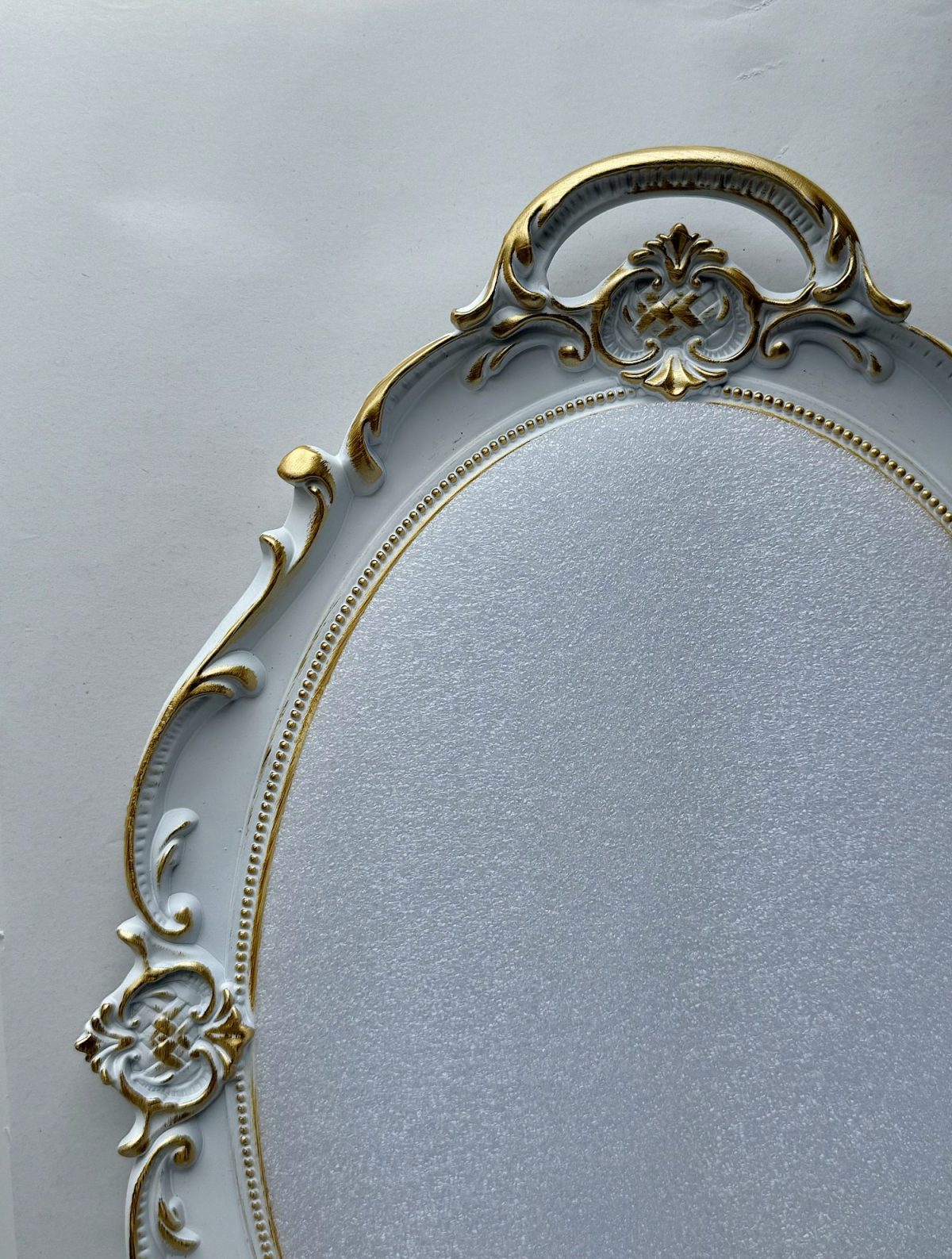 Ornate oval mirror frame with gold detailing and decorative floral accents against a plain light background.