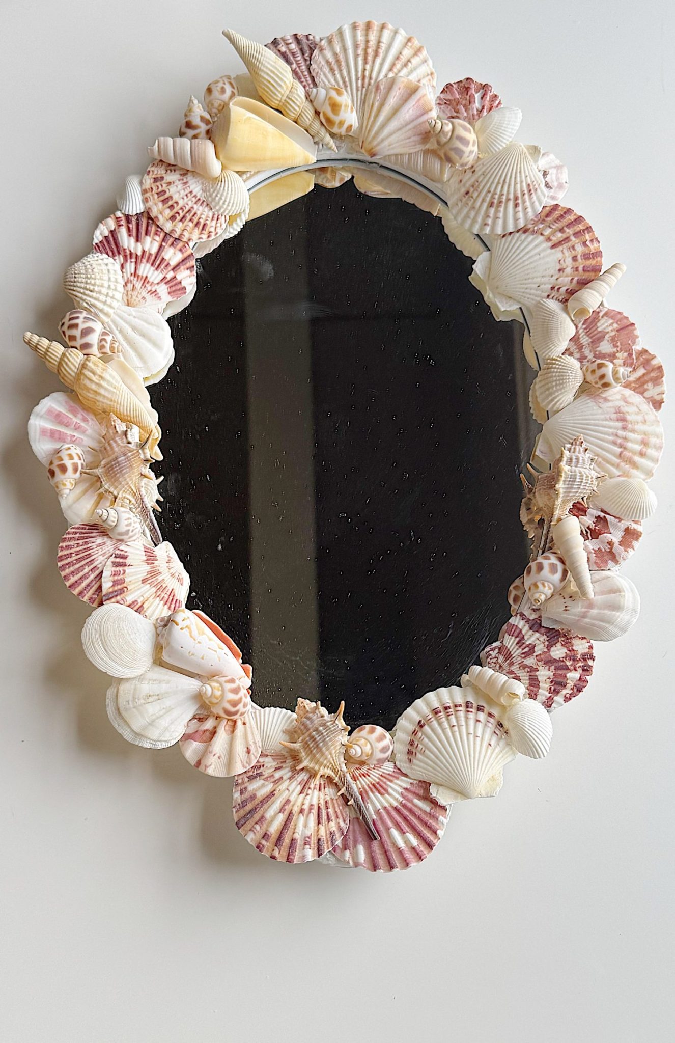 Oval mirror with a frame decorated with various seashells in shades of white, pink, and beige, hanging on a plain white wall.