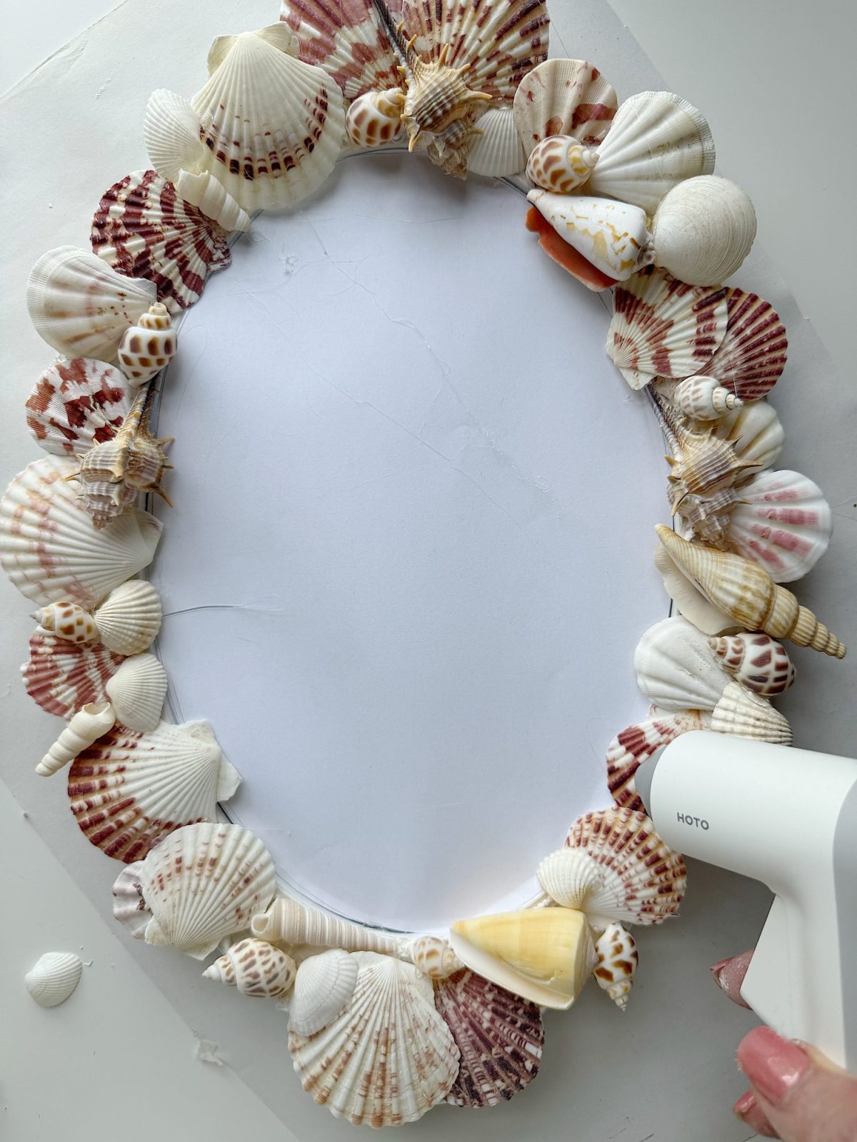 An oval-shaped frame is being decorated with assorted seashells using a hot glue gun.