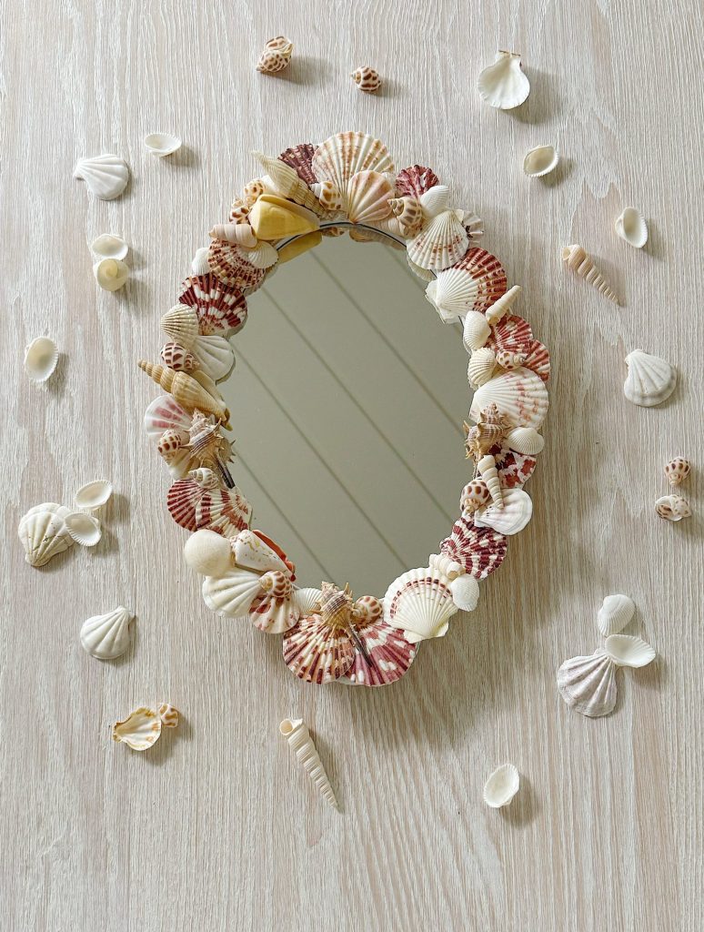 Oval mirror with a frame decorated with assorted seashells, surrounded by additional loose seashells on a light wooden surface.
