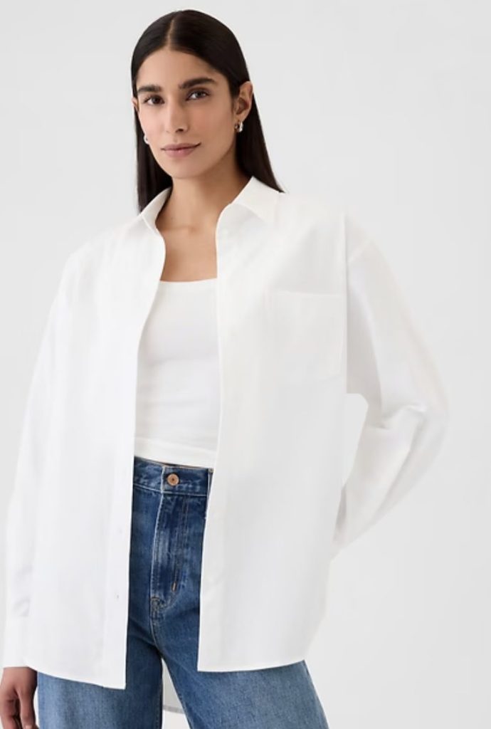 A person wearing a white button-up shirt over a white top with blue jeans, standing against a plain light background.