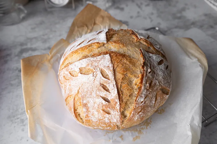 A round loaf of artisan bread with a golden crust and decorative scoring, resting on parchment paper on a metal cooling rack.