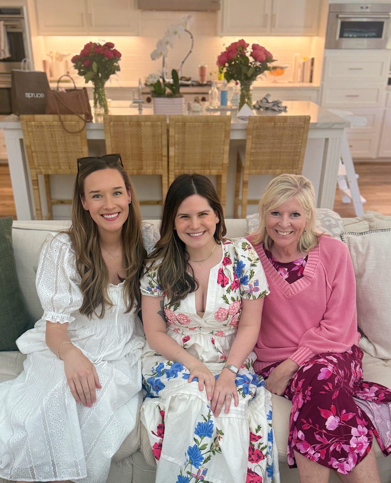 Three women are sitting on a couch in front of a kitchen, smiling at the camera. They are wearing spring dresses and the kitchen has flowers and bags on the counter.