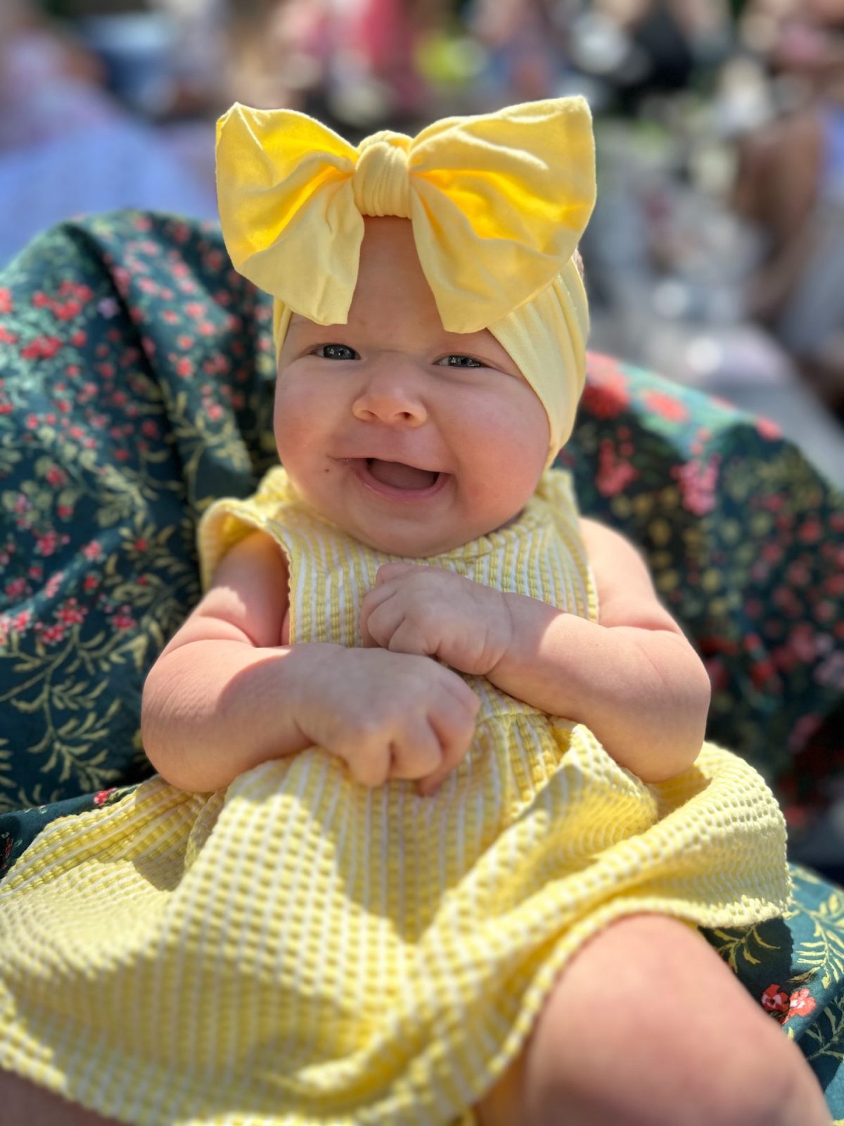Smiling baby wearing a yellow dress and large yellow bow headband, sitting on a floral-patterned blanket outdoors.