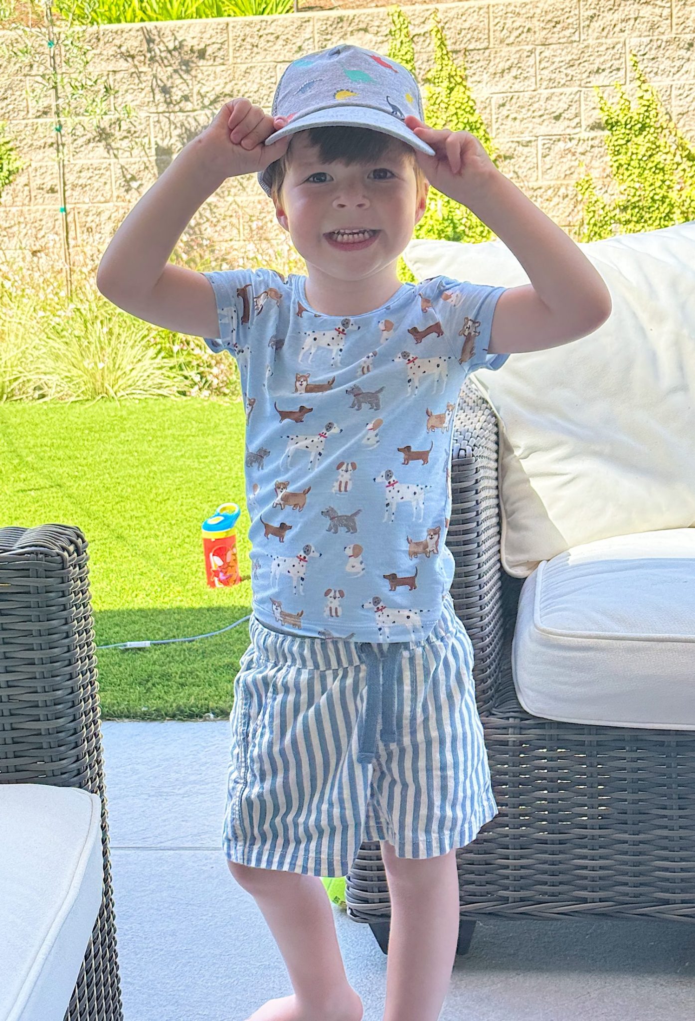 Young child standing on a patio, smiling and holding the brim of a cap, wearing a dog-patterned shirt and striped shorts, with outdoor furniture and green lawn in the background.