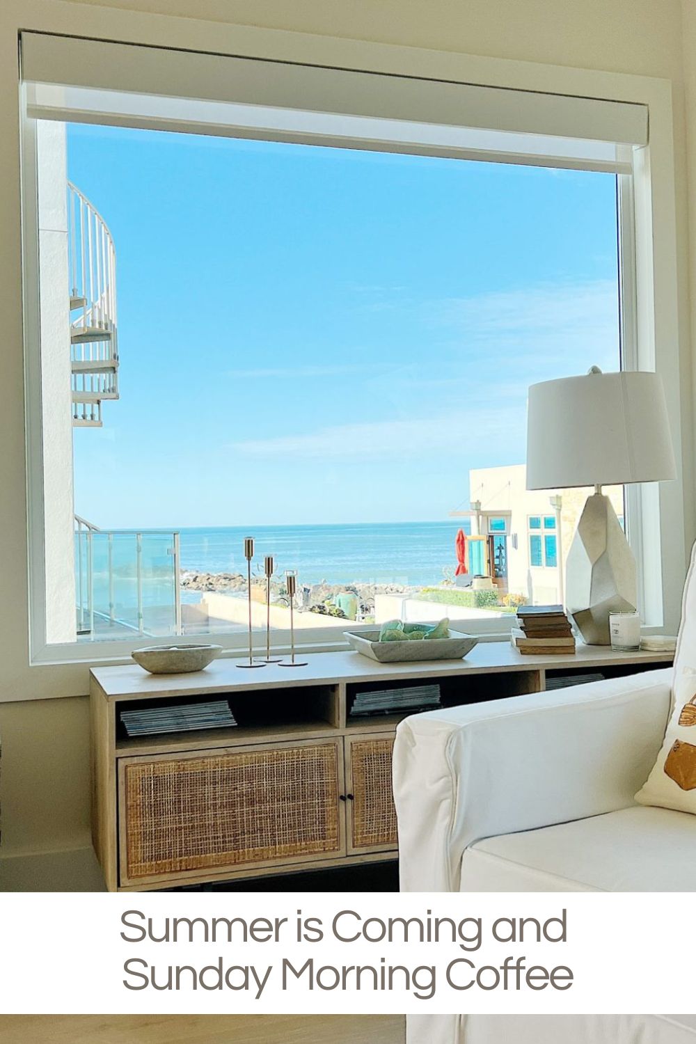 A bright living room with a large window overlooking the ocean, a spiral staircase outside, and a coffee table with decor and books in the foreground.