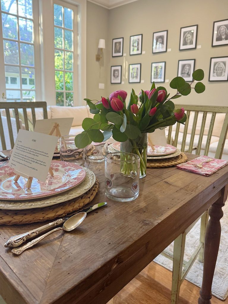 A wooden dining table set for two with floral plates, cutlery, glasses, a vase of pink tulips, and framed portraits on the wall in a bright room with large windows.