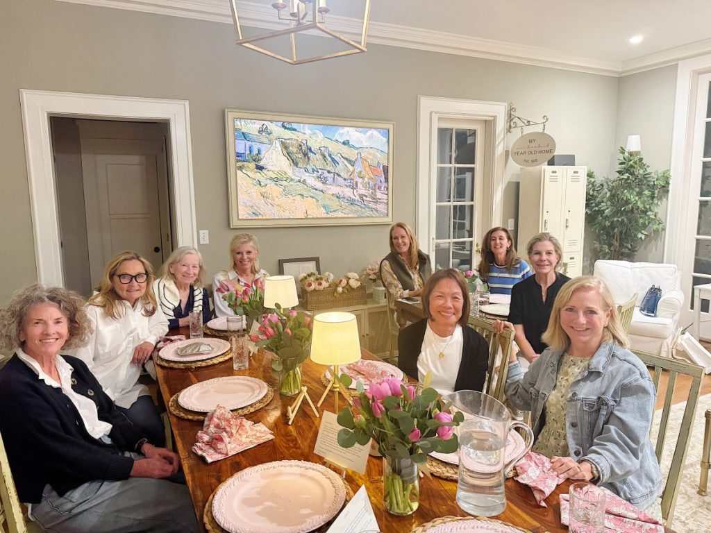 Nine women sit around a dining table set with pink plates, napkins, and floral centerpieces in a well-lit, decorated room.