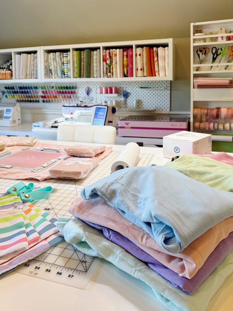A sewing room with folded pastel fabrics, striped fabric, cutting tools, a sewing machine, and shelves stocked with fabric rolls and supplies.