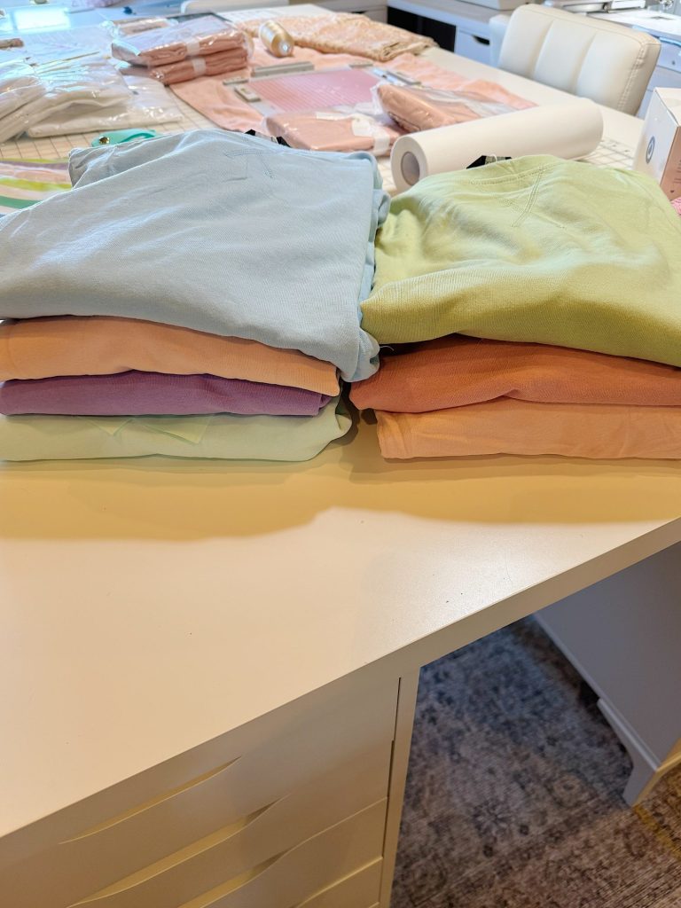Two stacks of folded pastel-colored clothes are placed on a white table, with more folded items and supplies in the background.