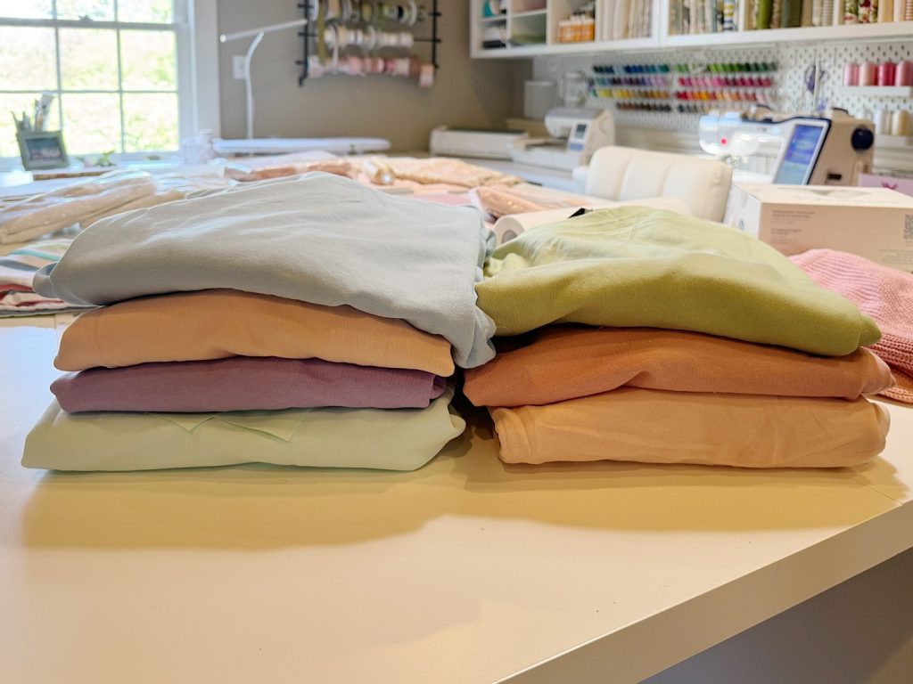 Two stacks of neatly folded pastel-colored fabric are placed on a white table in a well-lit craft room with sewing supplies in the background.
