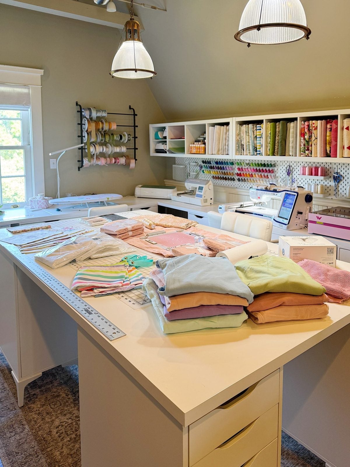 A well-organized sewing room with folding tables, sewing machines, fabric stacks, ribbons, and neatly arranged supplies on shelves and racks.