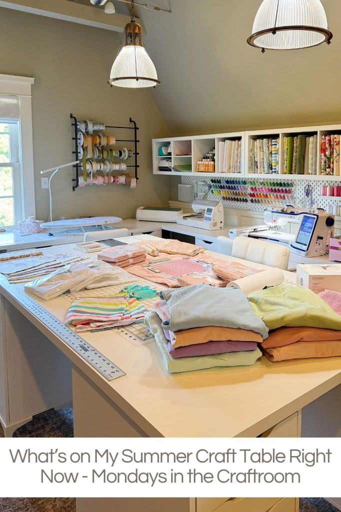 A craft room with organized shelves of fabric and ribbons, a sewing machine, folded textiles, and crafting tools on a large white table under pendant lights.