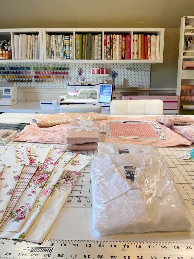 A sewing workspace with folded fabrics, a sewing machine, rulers on a grid cutting mat, and shelves filled with fabric and sewing supplies.