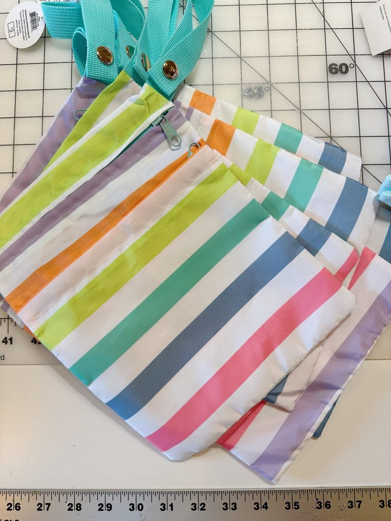 Three fabric pouches with colorful horizontal stripes and teal straps are laid out on a gridded cutting mat with measuring rulers.