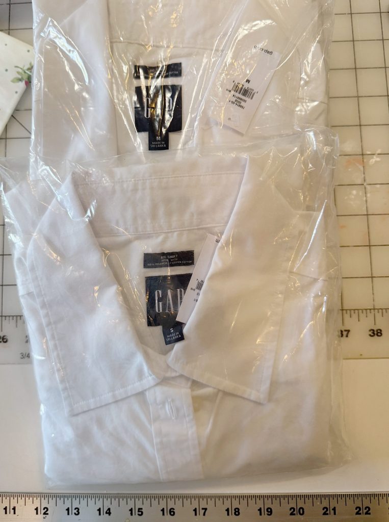 Two white collared GAP shirts, folded and packaged in clear plastic, are placed on a grid cutting mat with inch measurements along the edges.
