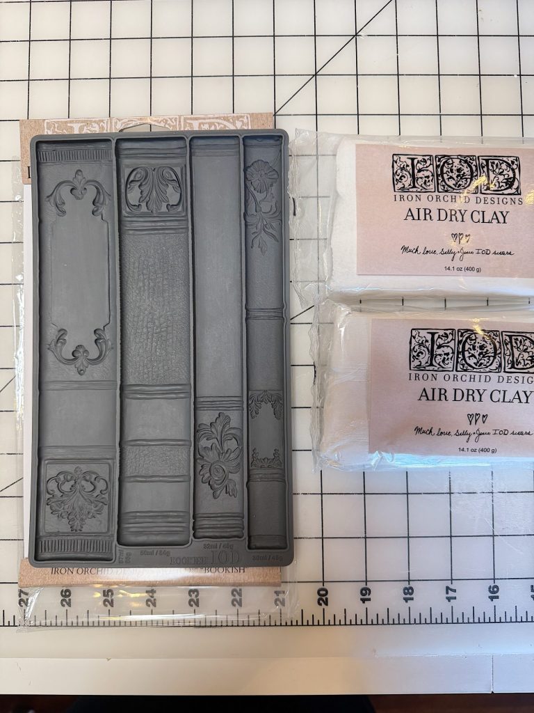 Rubber book spine mold and two packs of Iron Orchid Designs air dry clay are placed on a grid cutting mat.