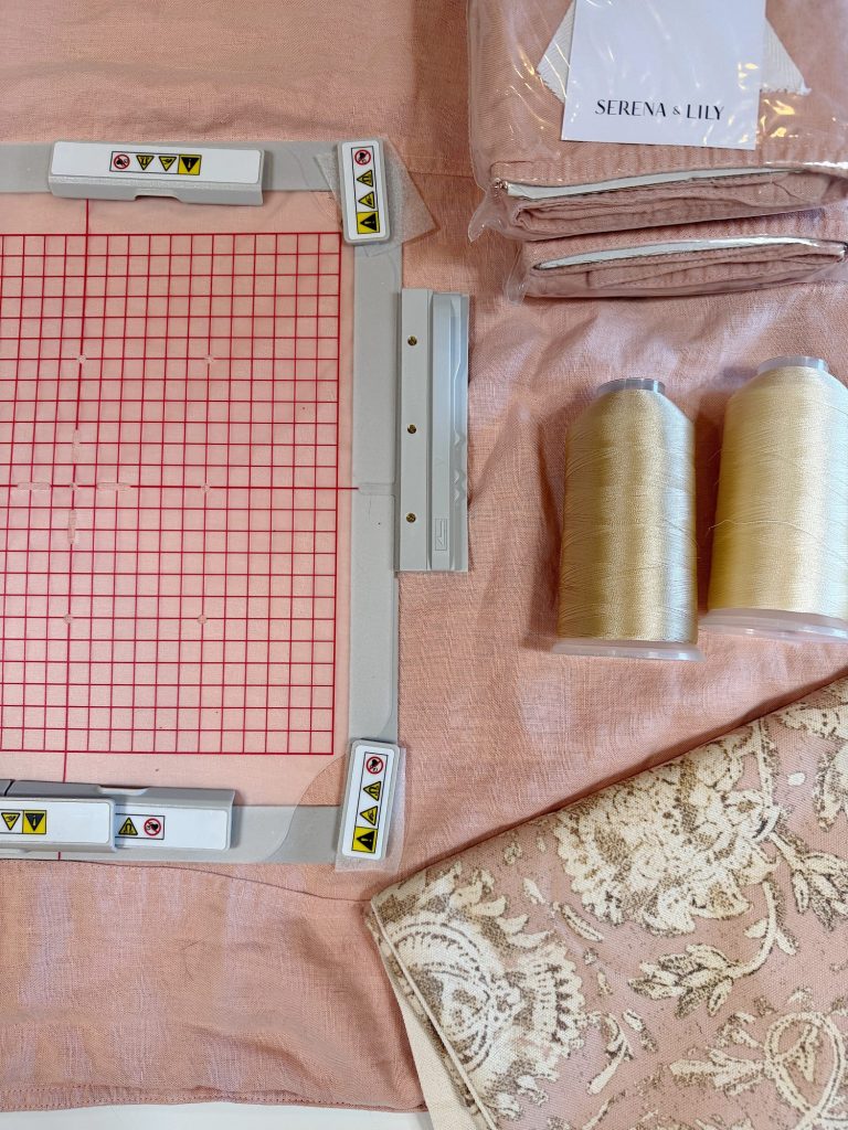 Assorted sewing supplies including a gridded embroidery hoop, two spools of beige thread, folded pink fabric, a Serena & Lily label, and a piece of patterned fabric.