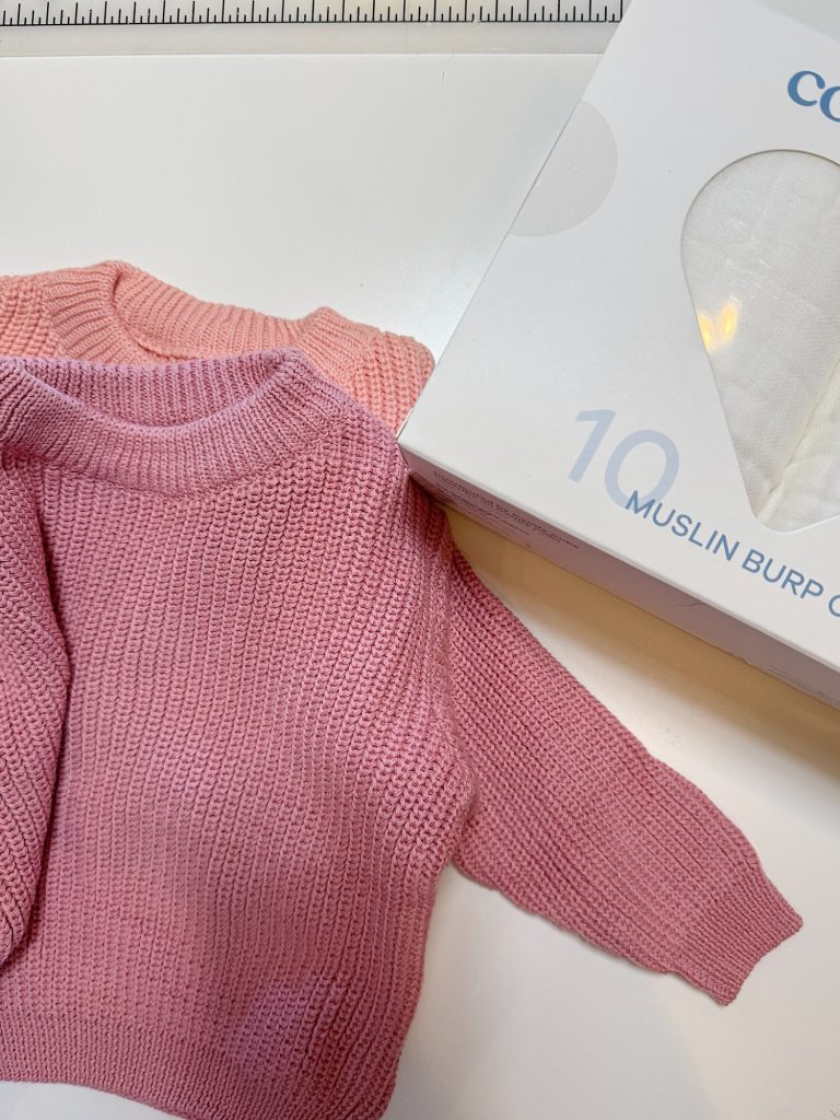 Two pink knitted baby sweaters next to a white box labeled "10 Muslin Burp Cloths" on a white surface.