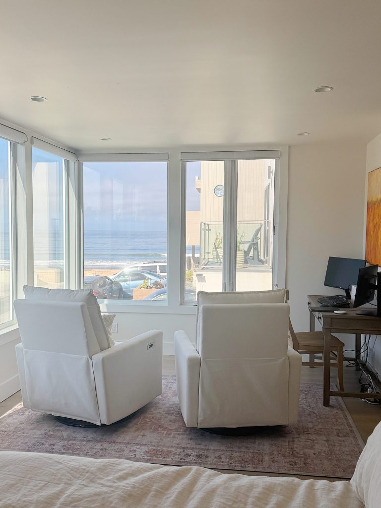 Two white armchairs face large windows with an ocean view; a desk with a computer is on the right side of the room.