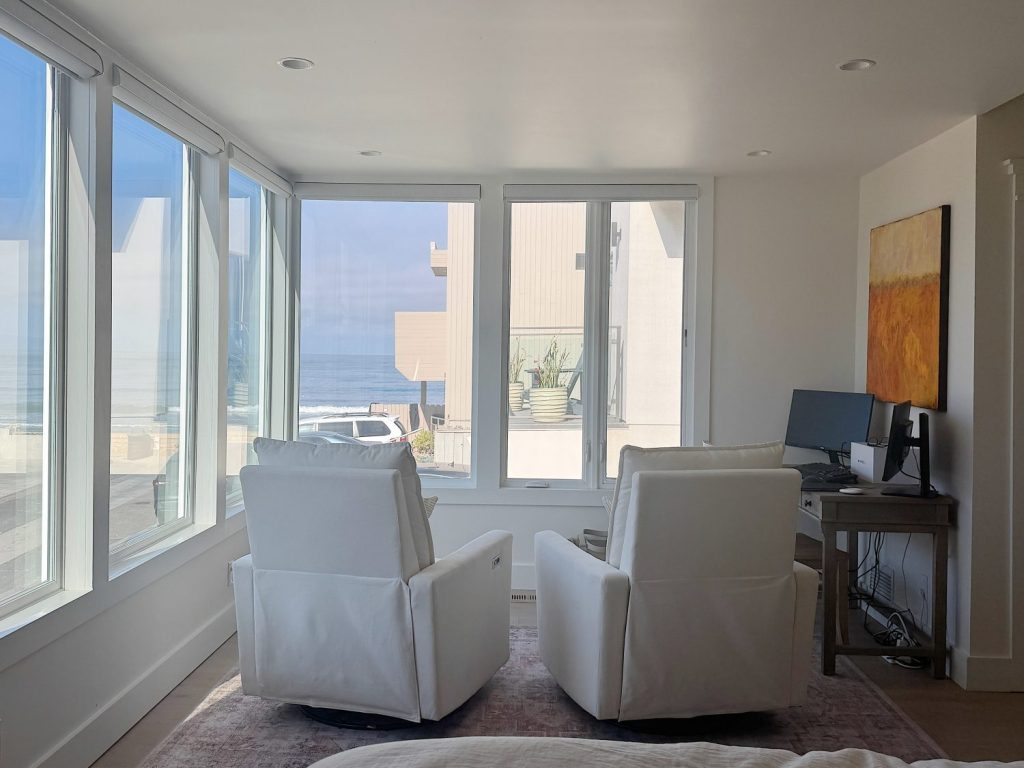 Two white armchairs face large windows with an ocean view. A small desk with a computer sits to the right, and a painting hangs on the wall above the desk.