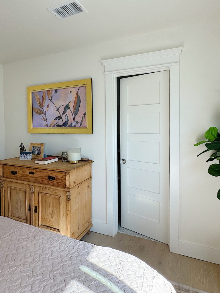 A wooden dresser with decor and framed photos sits against a white wall next to a partially open white door; a plant and a framed abstract art piece are also visible.
