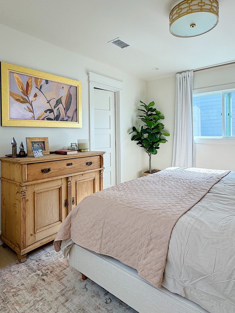A tidy bedroom with a beige quilted bed, wooden dresser, potted plant, framed artwork, and a window with white curtains.