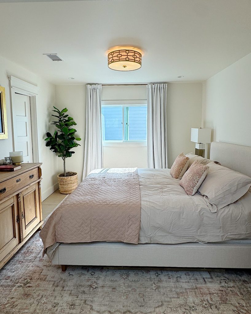 A neatly made bed with beige and pink bedding sits in a bright bedroom with a large window, a potted plant, a wooden dresser, and light-colored walls and curtains.