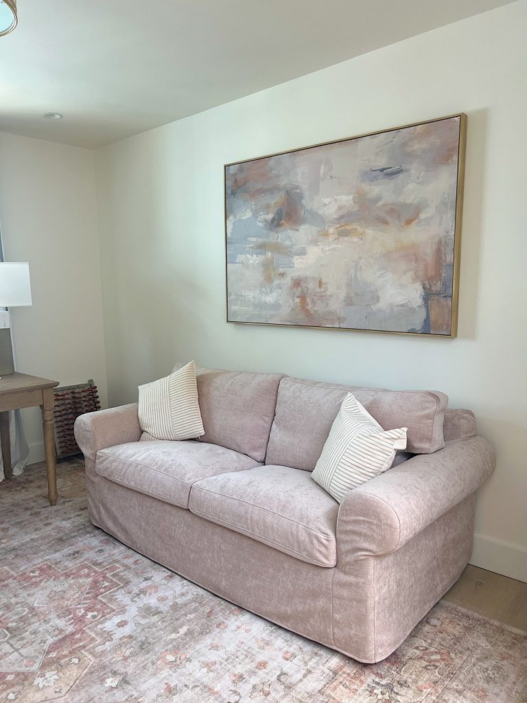 Beige sofa with two white striped pillows sits against a white wall beneath an abstract painting, with a patterned rug and wooden table nearby.