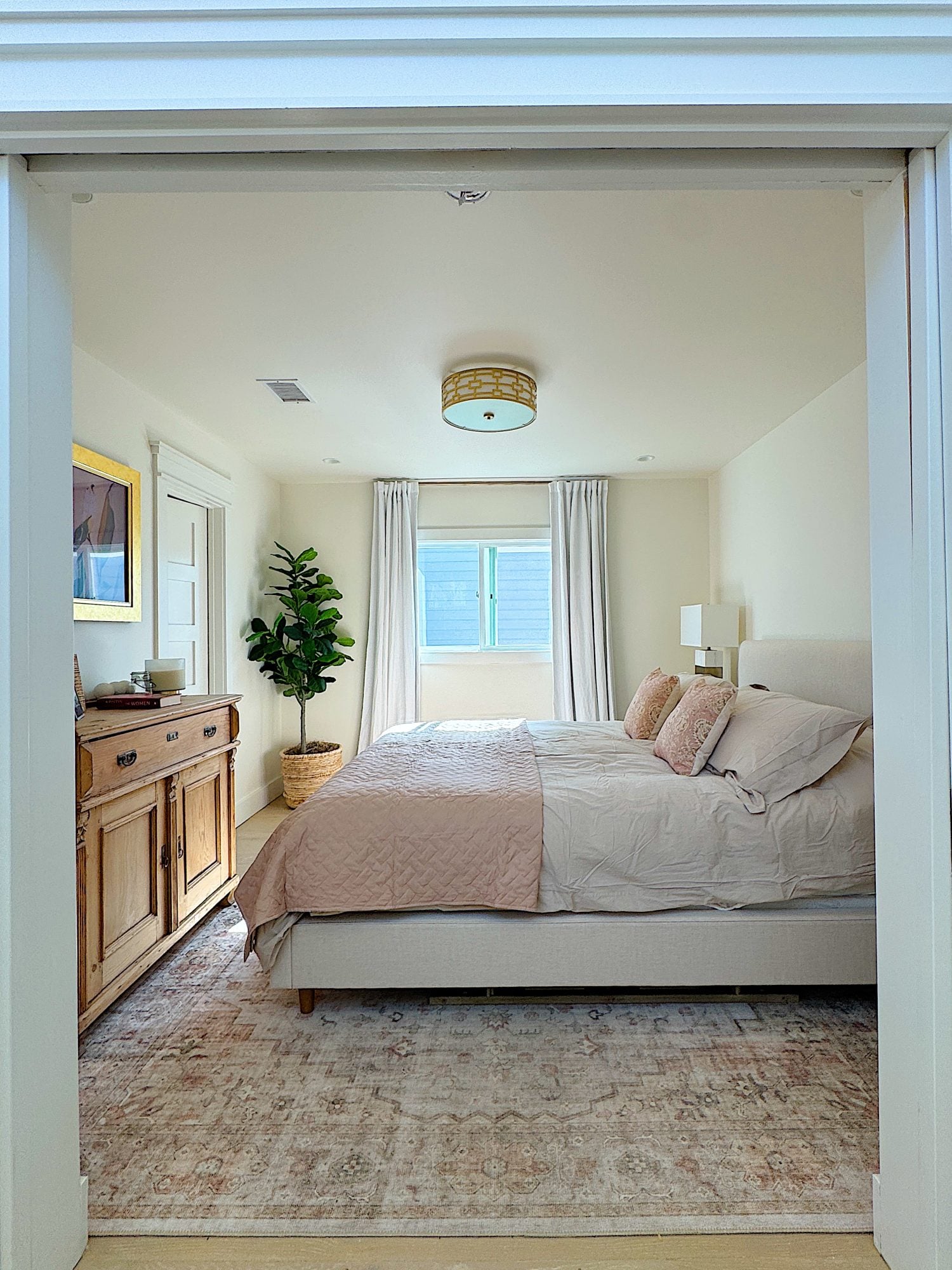 A bright bedroom with a large bed, light bedding, a wooden dresser, a potted plant, a window with curtains, and a ceiling light fixture.