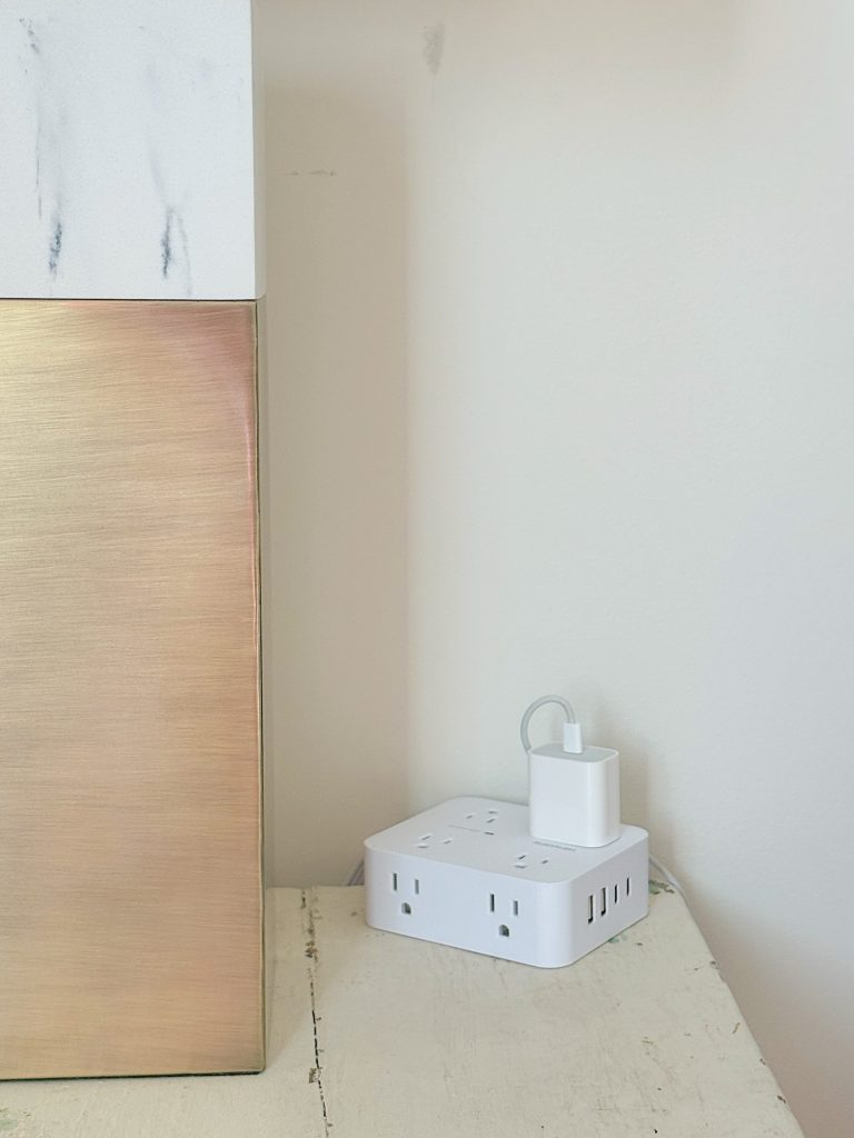 A white cube-shaped power strip with multiple outlets and a plugged-in charger sits on a light-colored surface next to a gold and marble decorative object against a white wall.