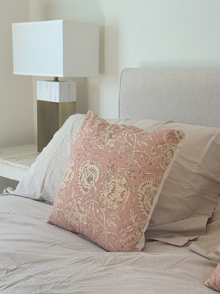 A bed with light beige sheets and a pink decorative pillow featuring a floral pattern, next to a modern white and gold table lamp on a bedside table.