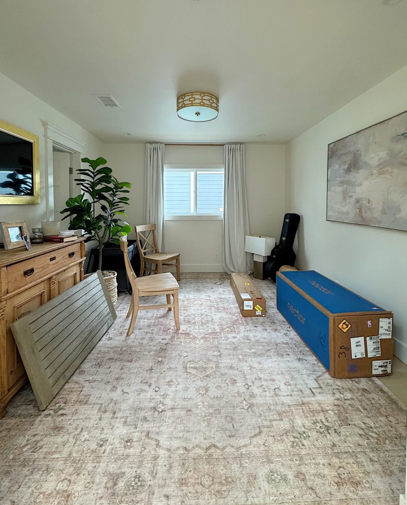 A room with light walls and a patterned rug, featuring a wooden cabinet, two chairs, boxed items, a large plant, a guitar case, and a window with white curtains.