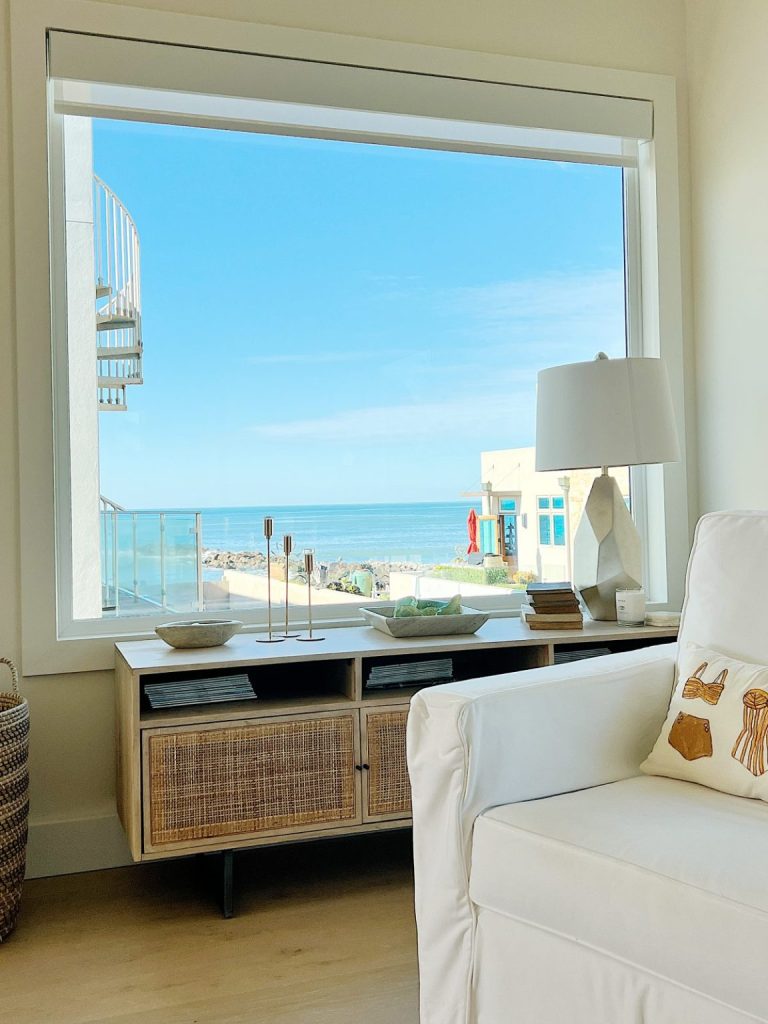 A large window in a bright room offers a view of the ocean, a spiral staircase, and a building outside. Inside, there&rsquo;s a white couch, lamp, and wooden console with decorative items.