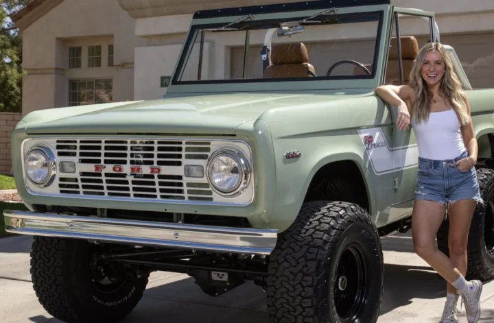 A woman in a white tank top and denim shorts stands smiling next to a vintage mint green Ford Bronco parked in a driveway.