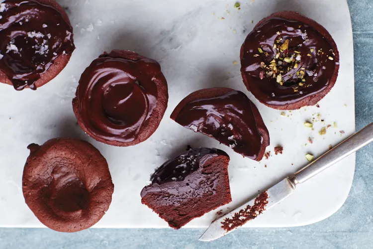 Five chocolate cupcakes with glossy chocolate frosting are arranged on a marble surface, with one cupcake cut in half and a knife with chocolate residue nearby.