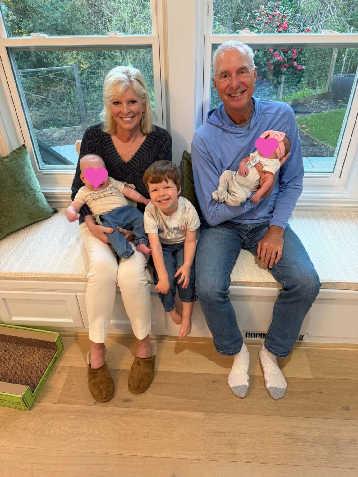 Two adults sit on a window bench, each holding a baby. A young child sits between them, smiling. The scene appears indoors with garden views outside the window.