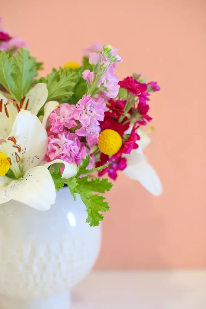 A white vase holds a bouquet of white lilies, pink and red flowers, yellow billy balls, and green leaves against a peach-colored background.