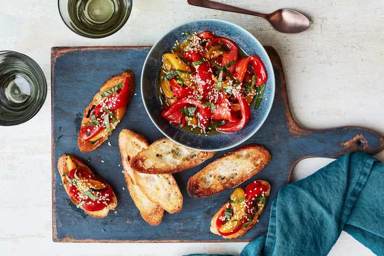 A bowl of tomato salad with sesame seeds and herbs, surrounded by toasted bread slices on a wooden board, with two glasses of white wine and a blue napkin nearby.