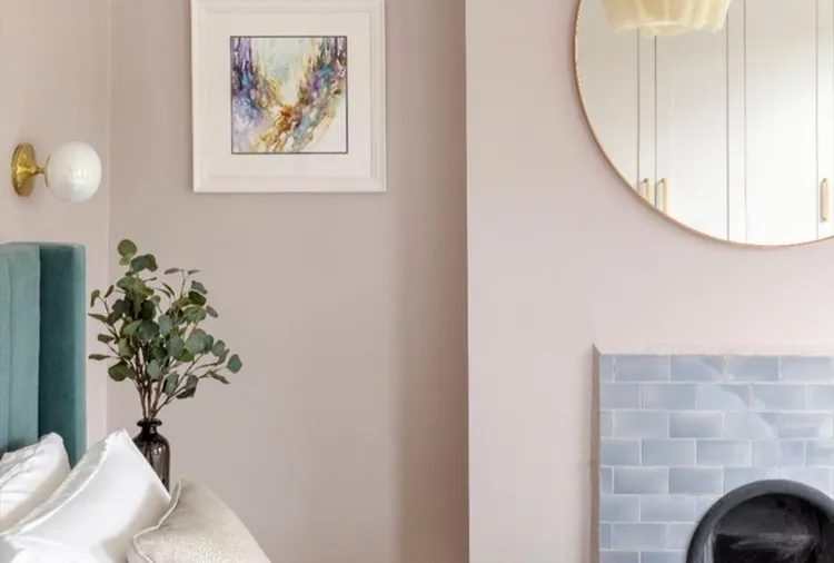Bedroom corner with a teal headboard, white bedding, a vase with greenery, abstract art on the wall, a round mirror, and a tiled fireplace.