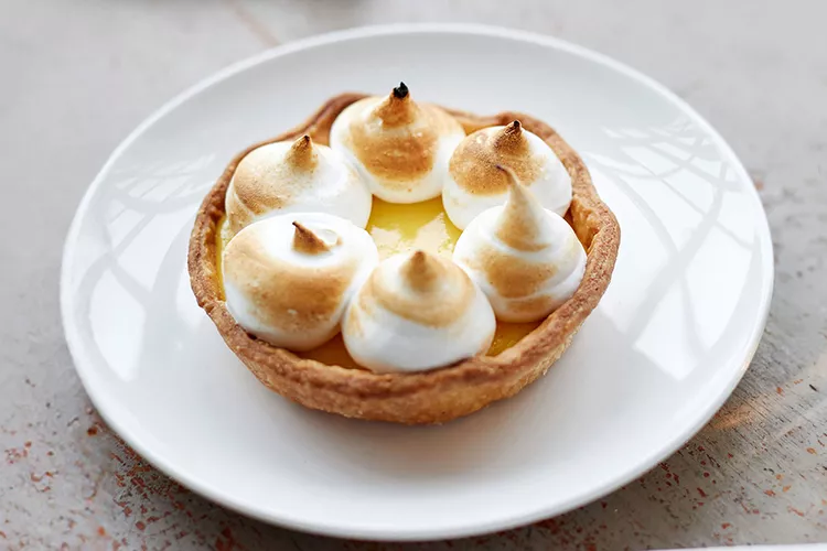 A lemon meringue tart with toasted meringue peaks sits on a white plate.