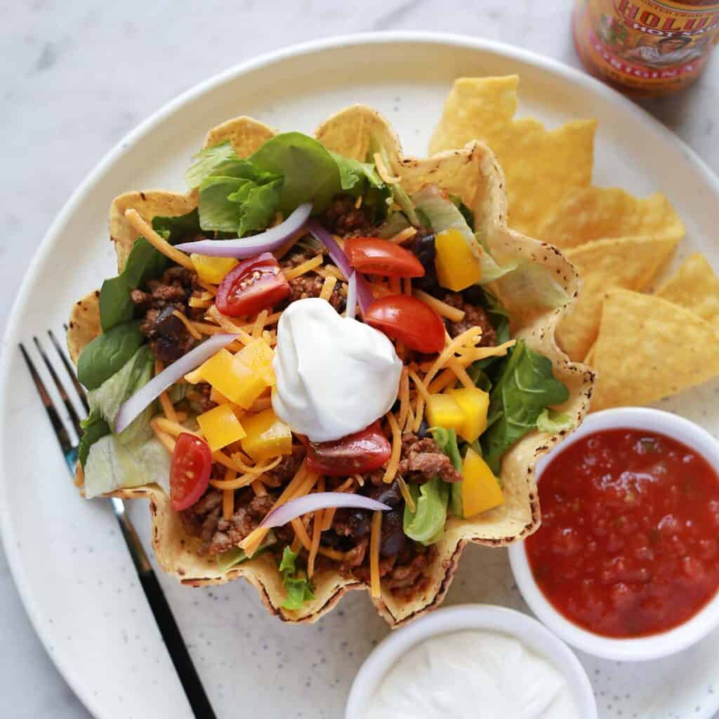 A taco salad in a tortilla bowl is topped with sour cream, shredded cheese, tomatoes, and onions, served with tortilla chips, salsa, and extra sour cream on a white plate.