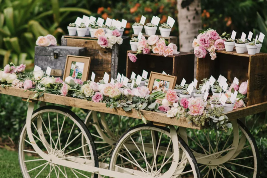 Wooden cart decorated with pink flowers, framed photos, potted plants, and cards, set outdoors in a garden setting.