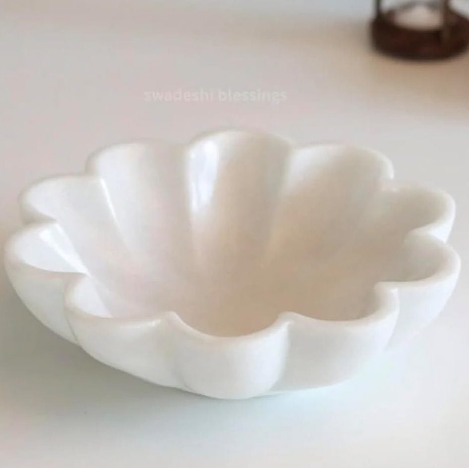 A white, scalloped-edge ceramic bowl, handcrafted with air dry clay, sits on a light surface with an hourglass, a blue patterned object, and a vase of flowers in the background.
