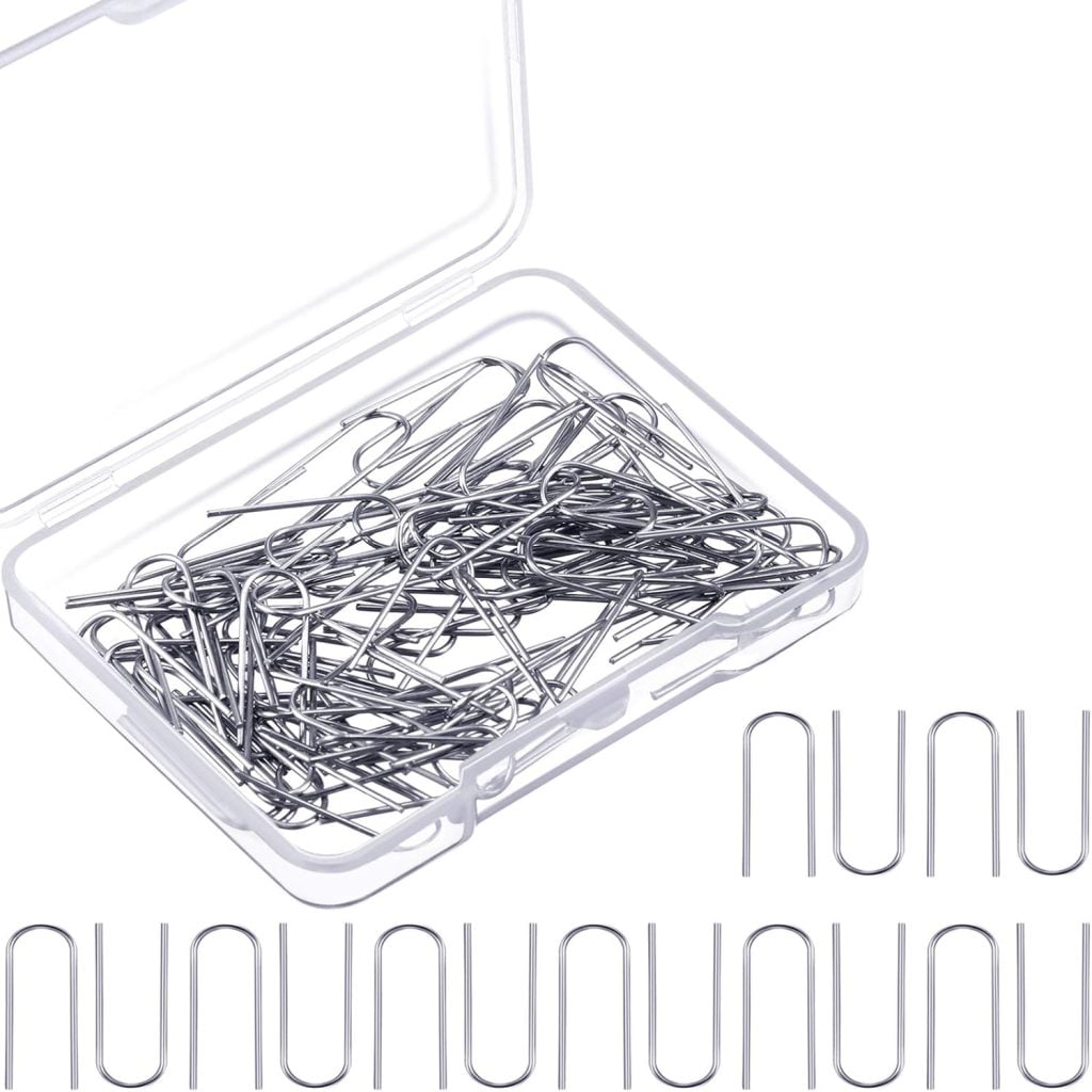A clear plastic box filled with silver U-shaped hair pins, displayed alongside delicate polymer clay charms. Several pins spell out "NUNU" in large letters using their unique shape.