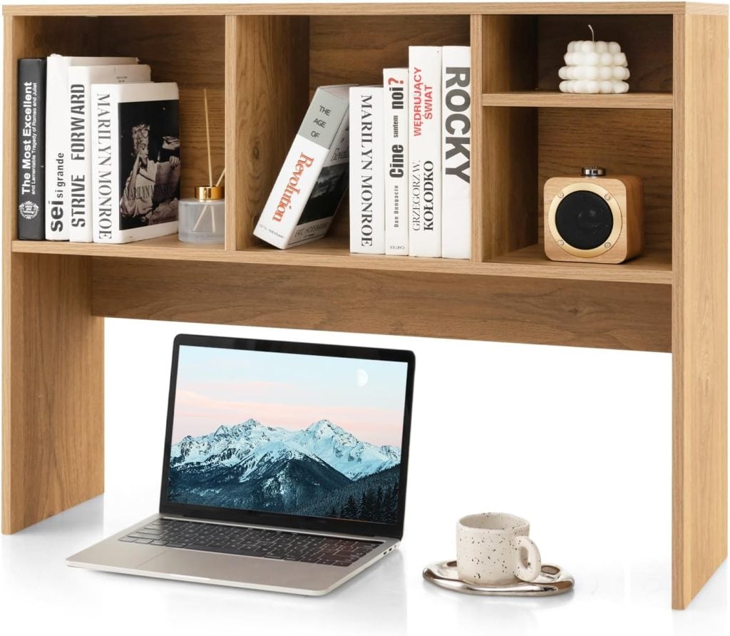 A wooden desk shelf holds books, a candle, a camera, and a perfume bottle. Below, a laptop with a mountain wallpaper and a cup with a saucer sit on the desk.