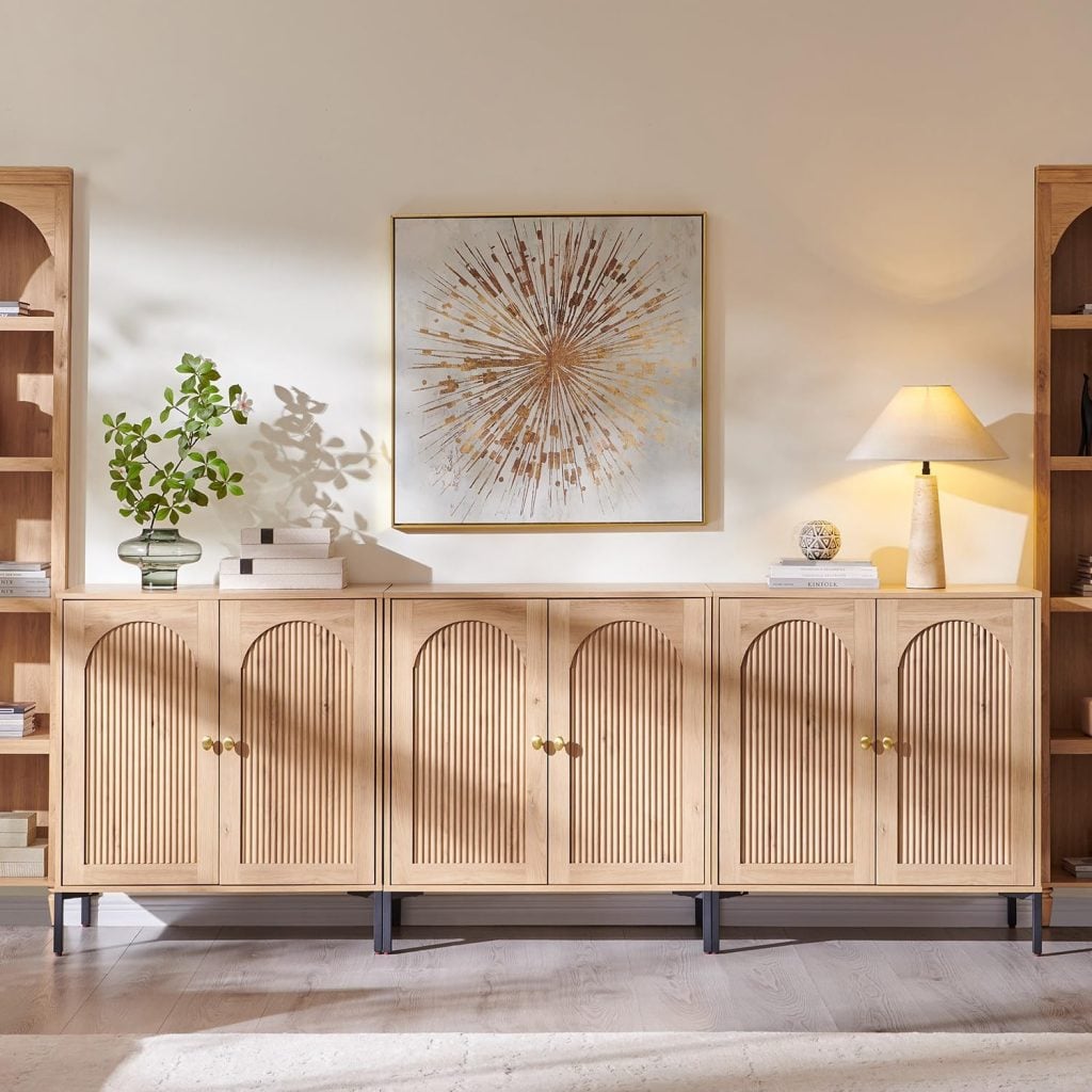 A wooden sideboard with arched cabinet doors stands against a wall, topped with a potted plant, a lamp, and decor, beneath a gold abstract art piece.