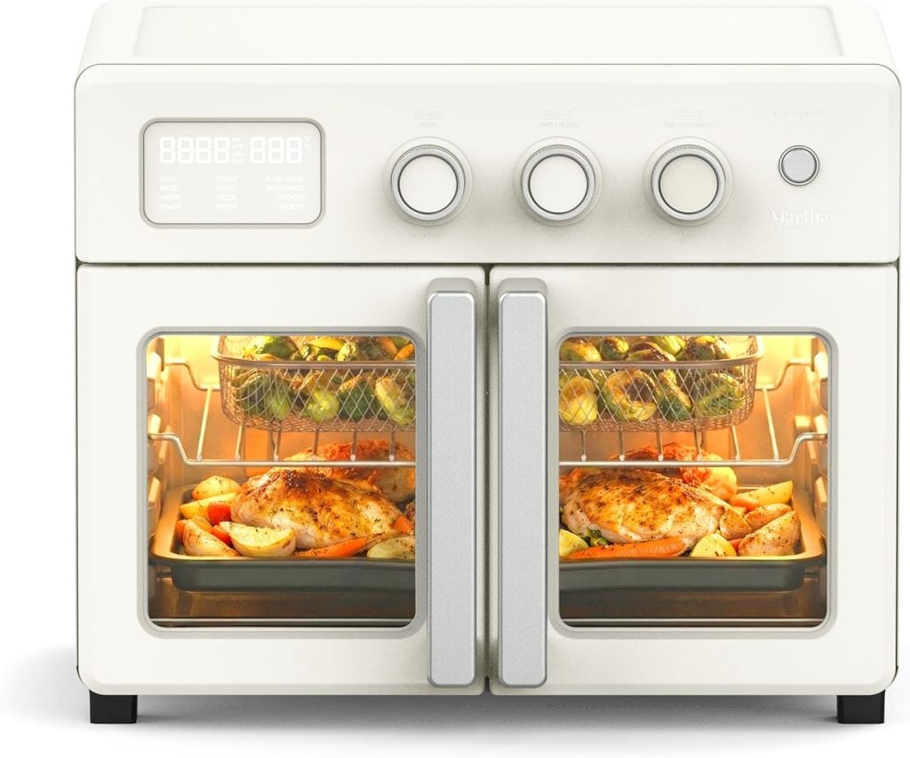 A countertop oven with a digital display and four dials, shown cooking a whole chicken with vegetables on the lower rack and roasted vegetables in a basket on the upper rack.