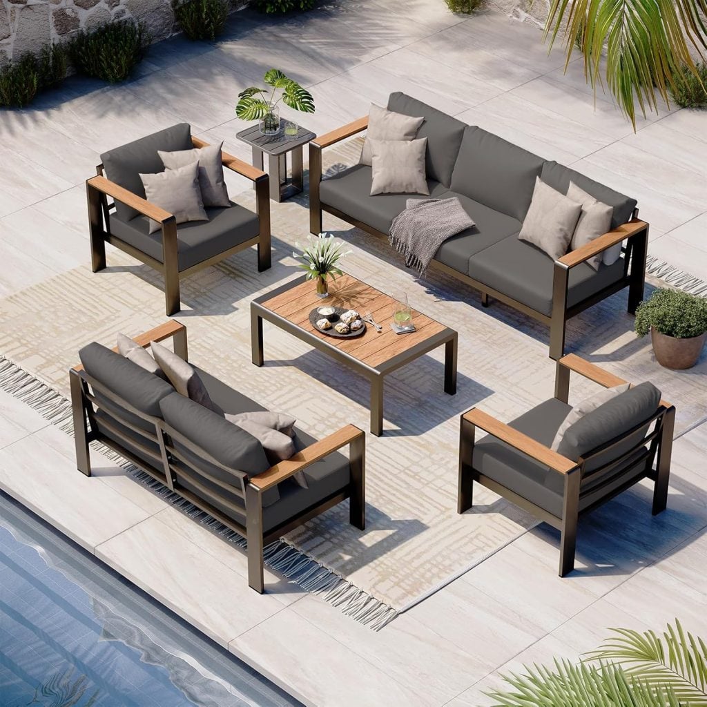 Modern outdoor patio set with two sofas, two armchairs, and a wooden coffee table arranged on a rug beside a pool, surrounded by plants.